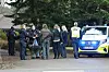 Politi og civile står ved en politibil på en stille villavej med træer i baggrunden.