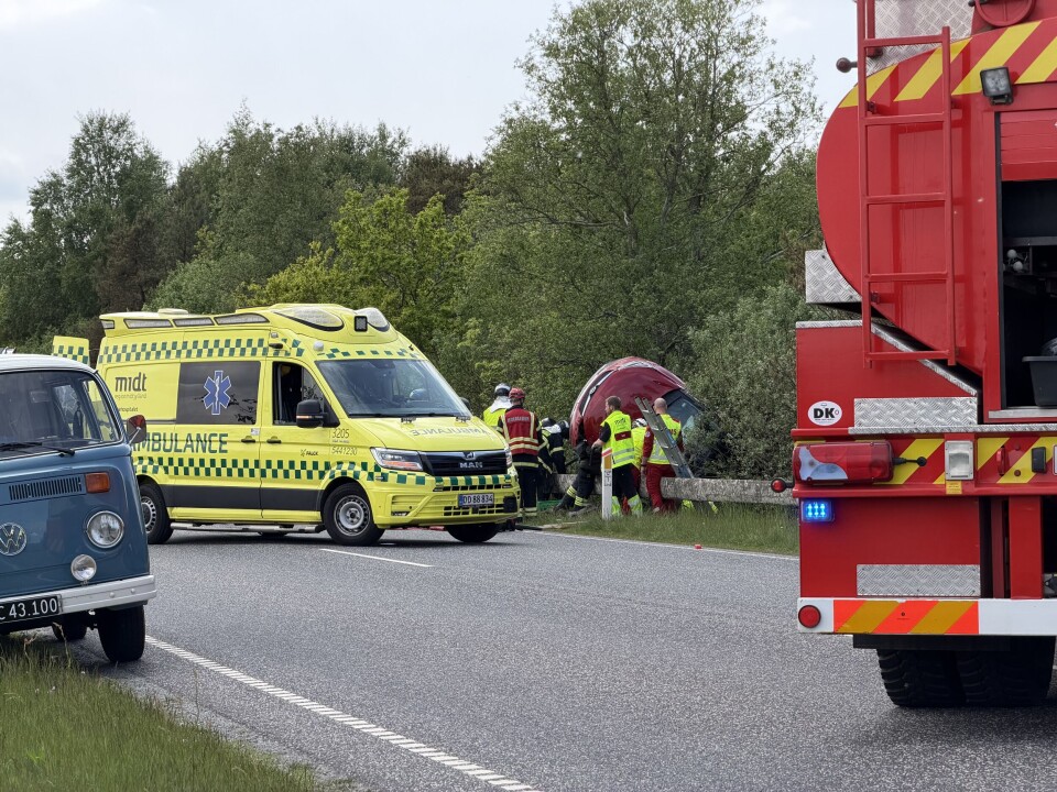 Redningsfolk ved en trafikulykke på en landevej med ambulance og stort rødt køretøj.