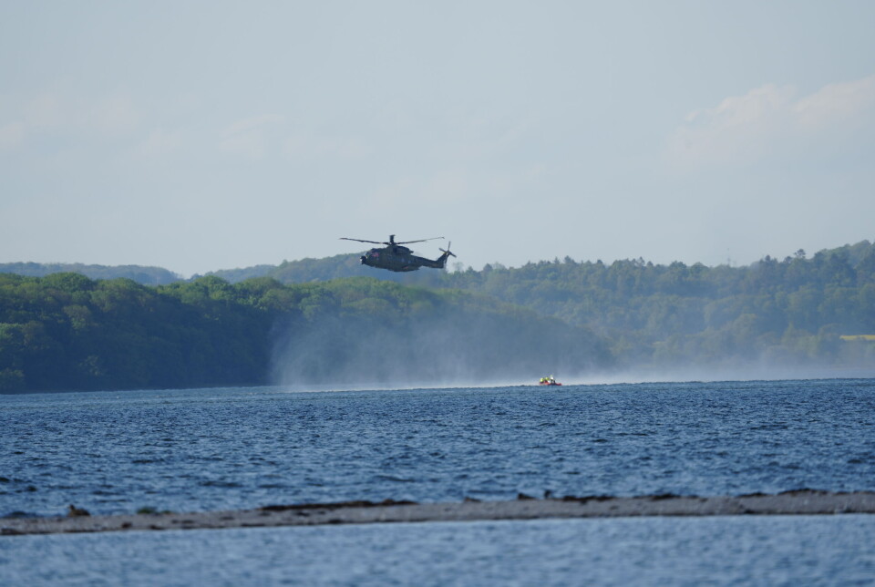 En helikopter svæver lavt over vandet med skov og bakker i baggrunden.