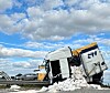 Hvid væltet lastbil med sække omkring sig ved en vej under blå himmel.